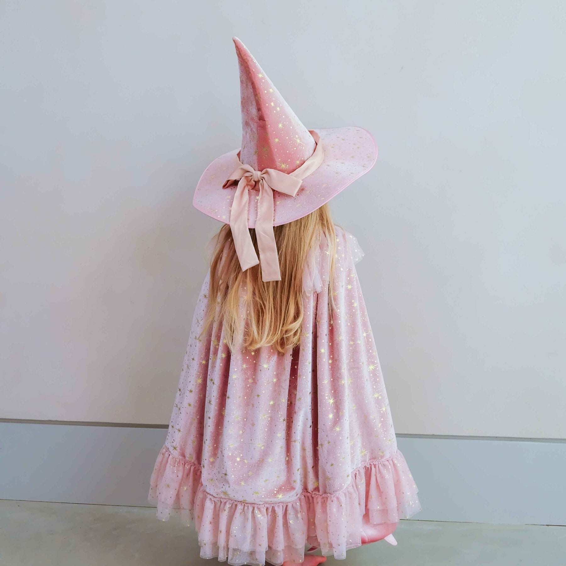 A young girl viewed from behind wearing a pink witch costume, featuring a tall pointed hat with a bow and a sparkly pink cape, standing against a plain light gray wall.