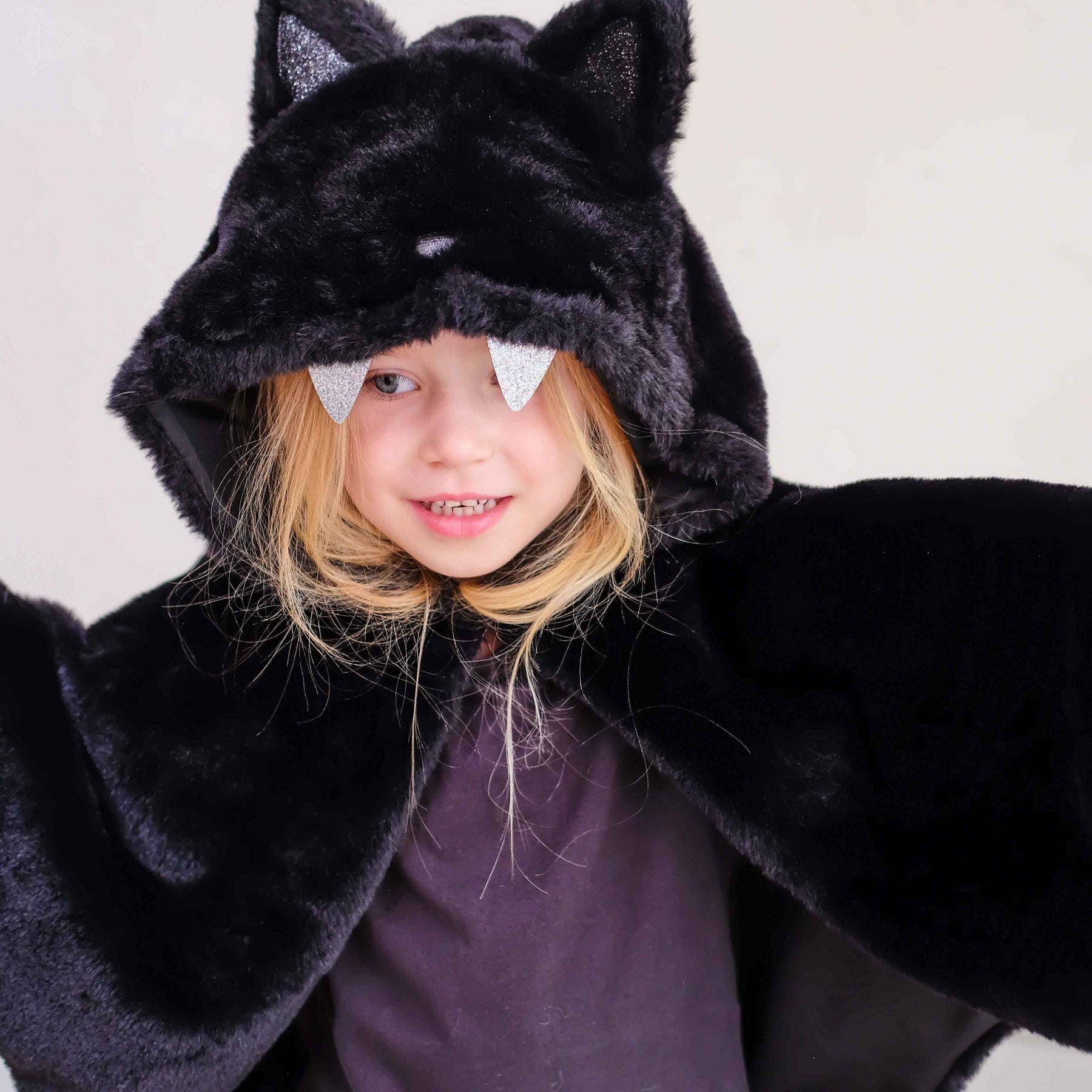 A child wearing a plush black bat costume hood with pointed ears, silver glitter bat wings attached to the hood, and a dark purple shirt underneath, photographed against a plain light background.