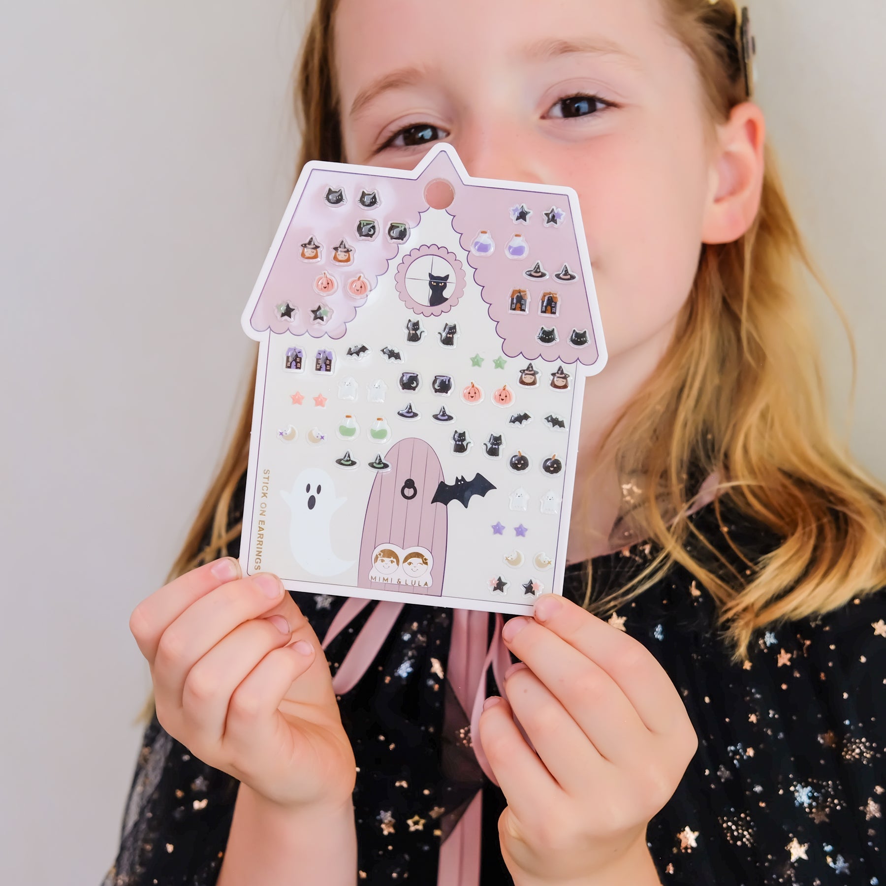 A young girl with blonde hair holds up a house-shaped sticker sheet decorated with Halloween-themed stickers like bats, ghosts, and pumpkins. She is wearing a black star-patterned top against a plain light gray background.