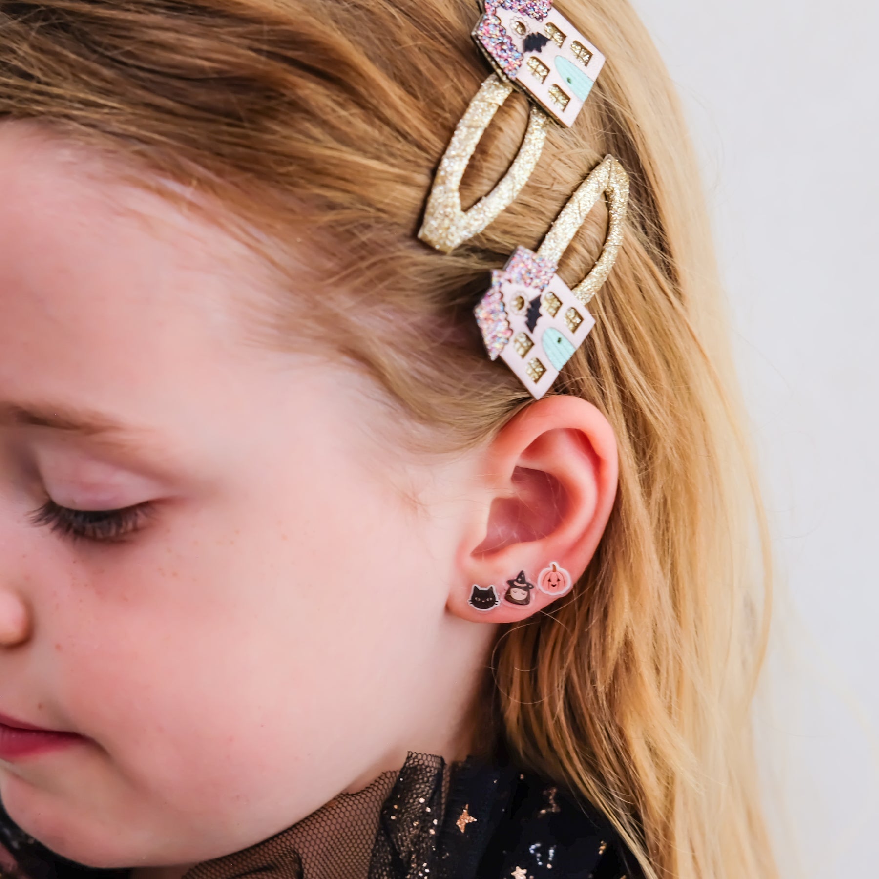 A close-up of a young girl's head and ear, showcasing two gold hair claw clips with colorful pastel house charms and matching small stud earrings.
