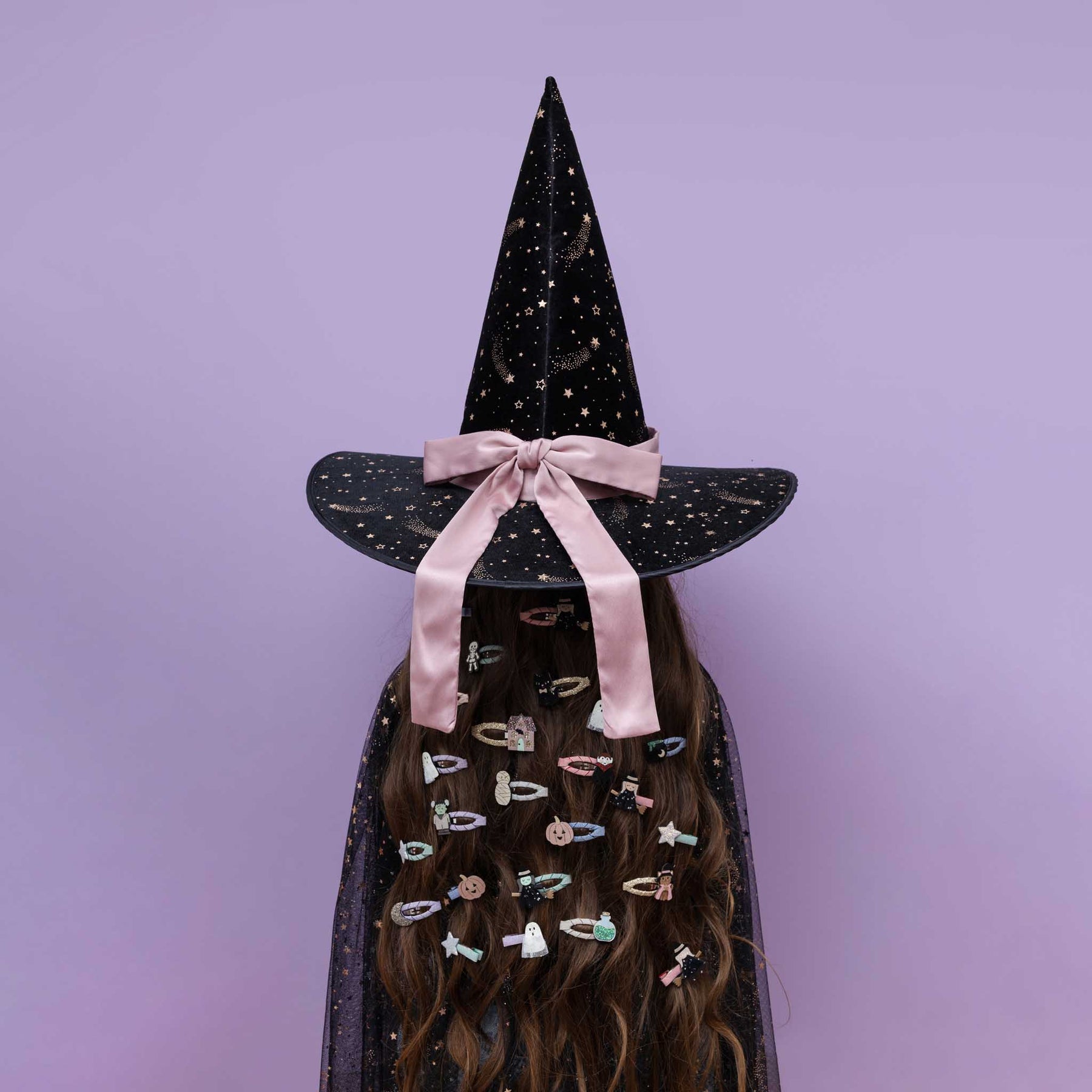 A person wearing a tall black witch hat with gold speckles, a pink satin bow, and a matching black cape covered in colorful hair clips, set against a solid lavender background.