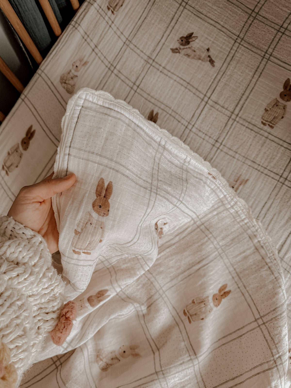 A close-up of a hand holding a cream-colored muslin blanket with a brown bunny print, set against a matching plaid sheet.