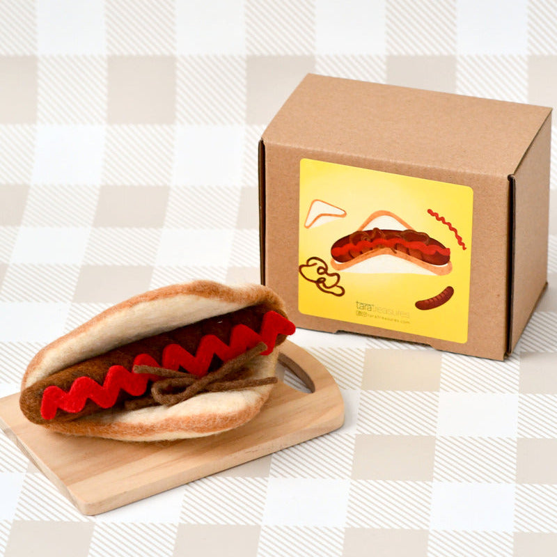 Hot dog-shaped product with bun and sausages on a wooden board, next to a cardboard box with a hot dog illustration.