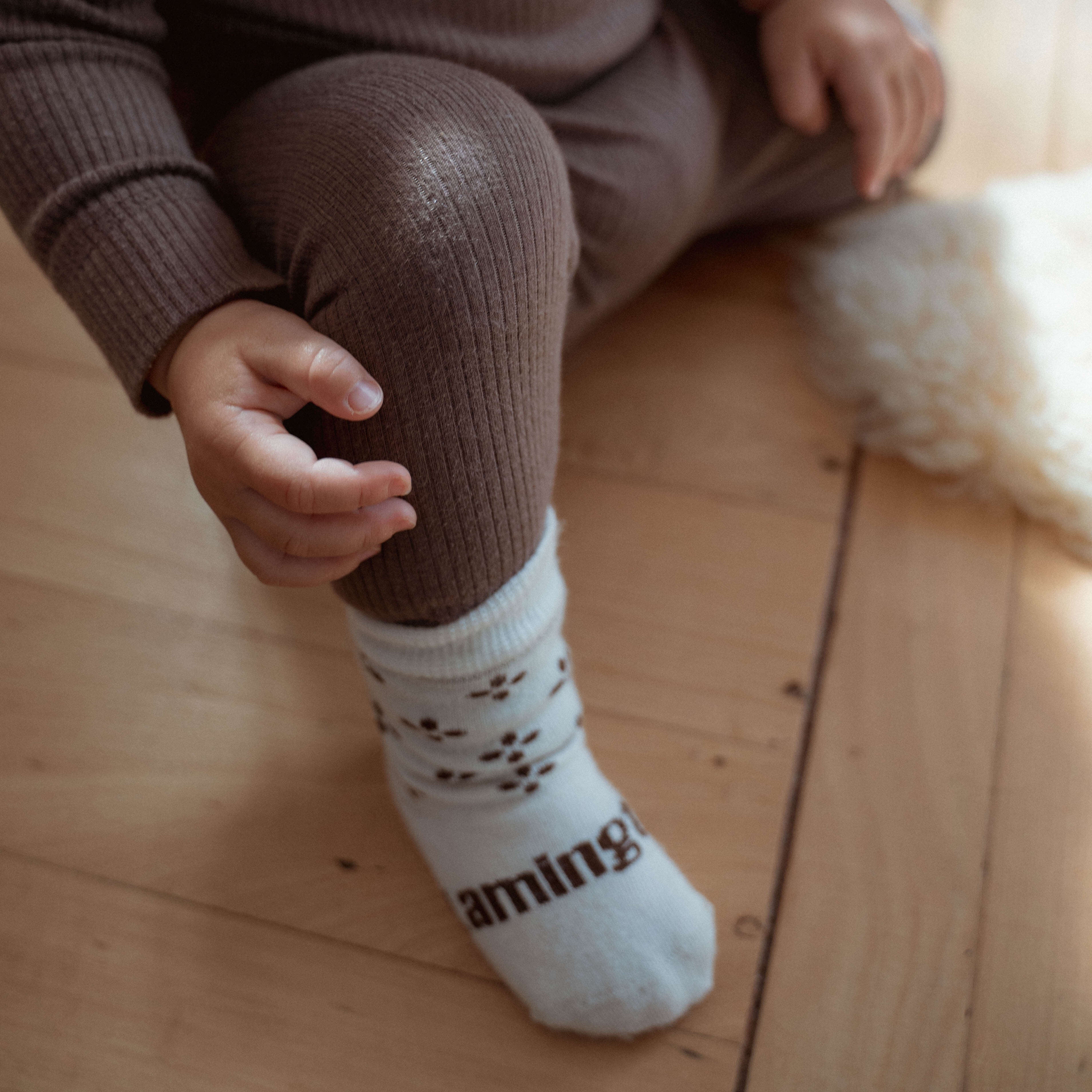 A close-up of a child wearing ribbed brown leggings and white socks with dark brown paw prints and the word 'aming' printed on the sole, sitting on a light wood floor.