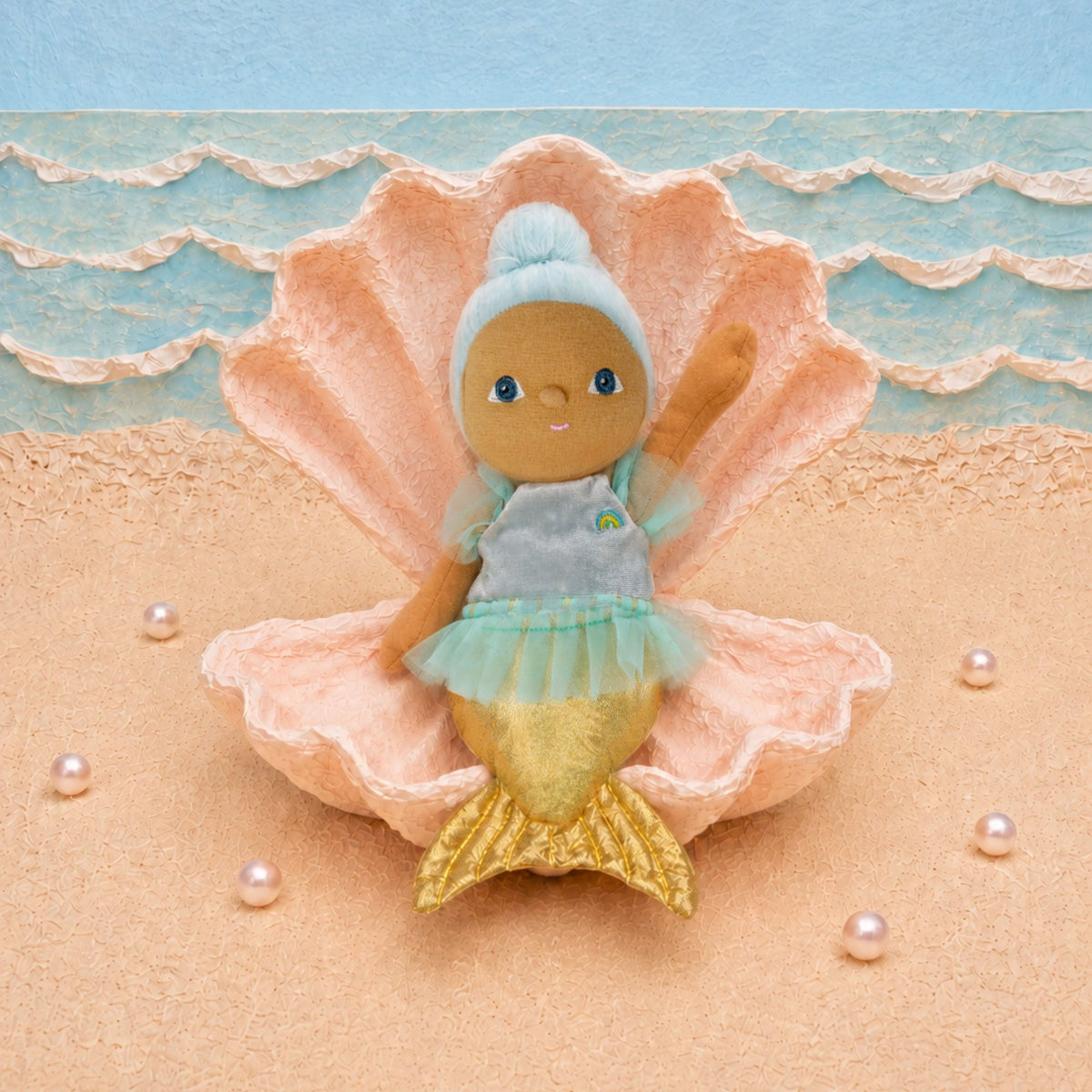 A plush mermaid doll with brown skin, blue hair, and a light blue and green dress sits inside a large pink seashell on a sandy beach set with blue ocean waves and pearl-like beads.