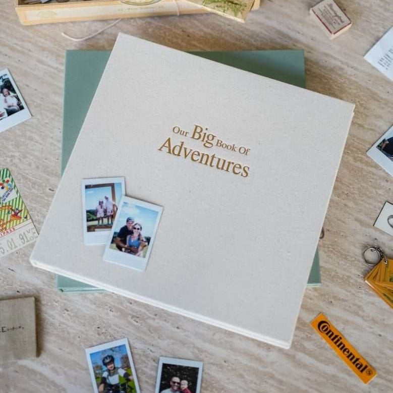 A square photo album titled 'Our Big Book Of Adventures' lies open on a light wood surface, surrounded by Polaroid-style photos, a green folder, and small travel-themed props. The album cover is a warm off-white textured paper with gold lettering.