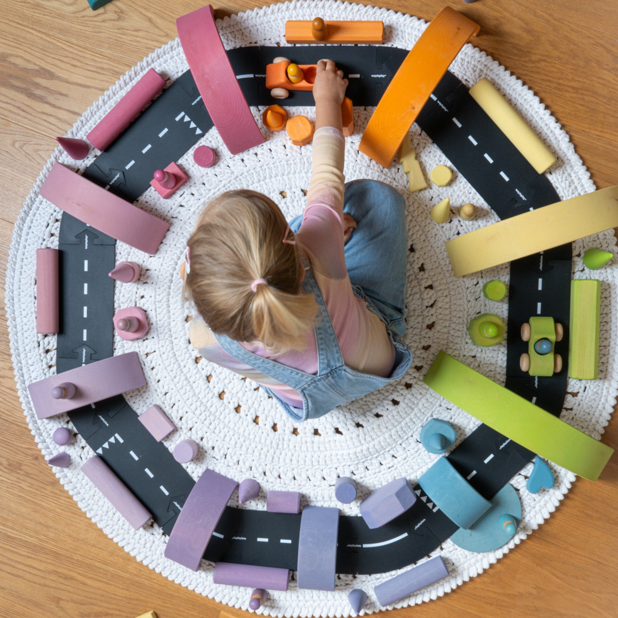 A child sits on a round white crocheted rug surrounded by a colorful wooden toy road set with black roads, pink and orange bridges, yellow and green sections, and small toy cars, on a wooden floor.