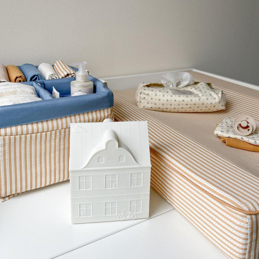 A styled baby changing table setup featuring a striped changing pad, a blue fabric diaper basket filled with baby items, a white ceramic house-shaped container, and a patterned tissue pouch.