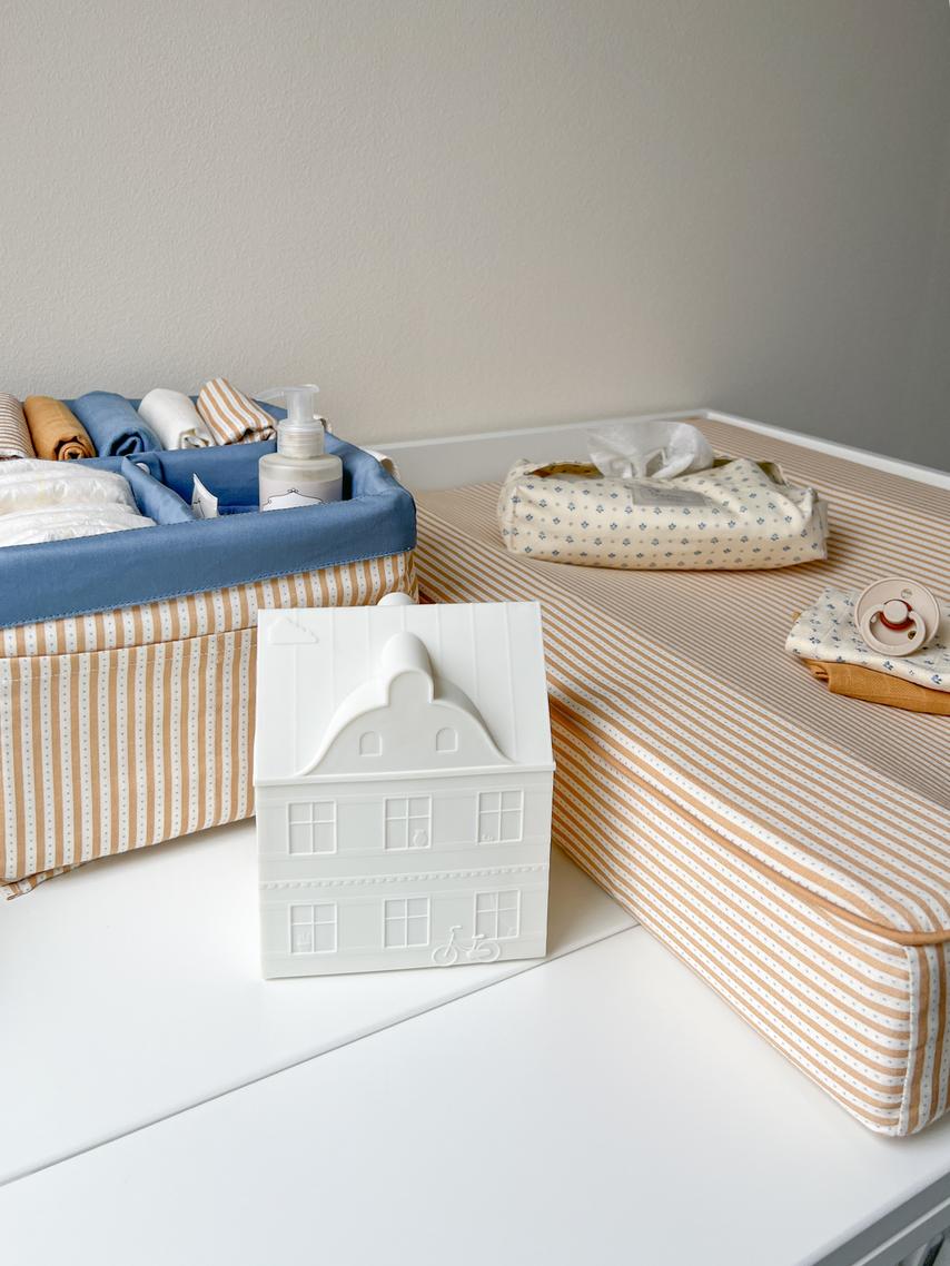 A styled baby changing table setup featuring a striped changing pad, a blue fabric diaper basket filled with baby items, a white ceramic house-shaped container, and a patterned tissue pouch.