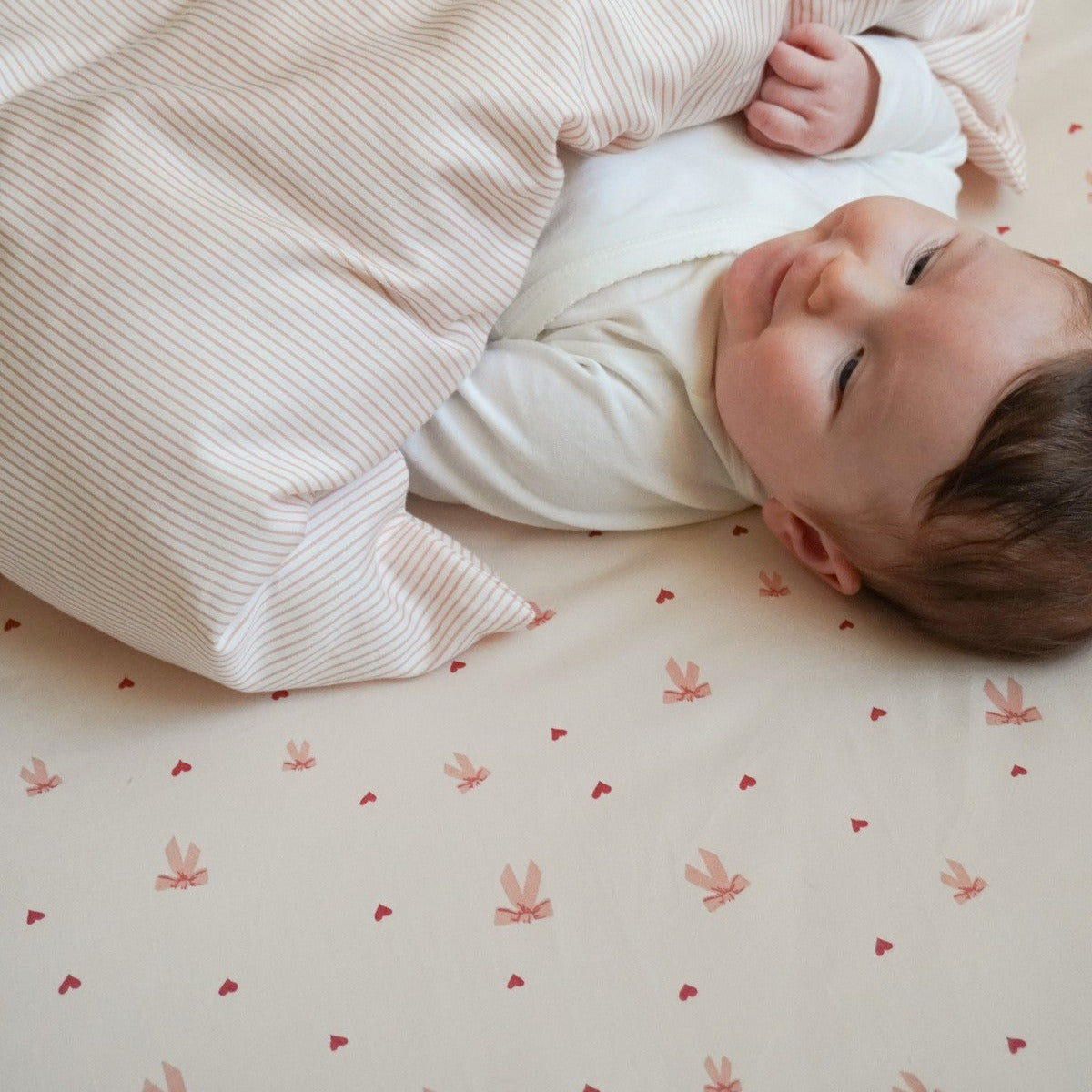 A baby lying on a cream-colored mattress, partially covered by a cream ribbed blanket. The mattress has a matching cream cover with a small pink bunny and heart pattern.