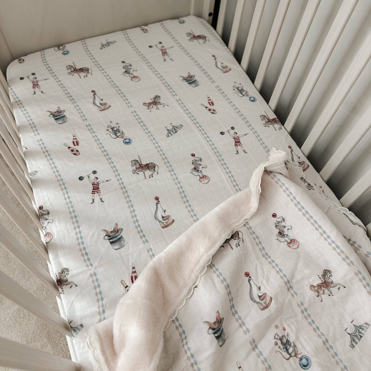 A baby crib mattress fitted sheet with a white background and a playful print of vintage carousel horses and riders in muted red, blue, and brown, with a matching plush cream blanket draped over the corner.