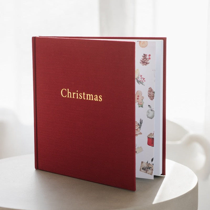A red hardcover book or album standing open on a round table, with the word 'Christmas' in gold on the cover and a white interior page decorated with small holiday illustrations.