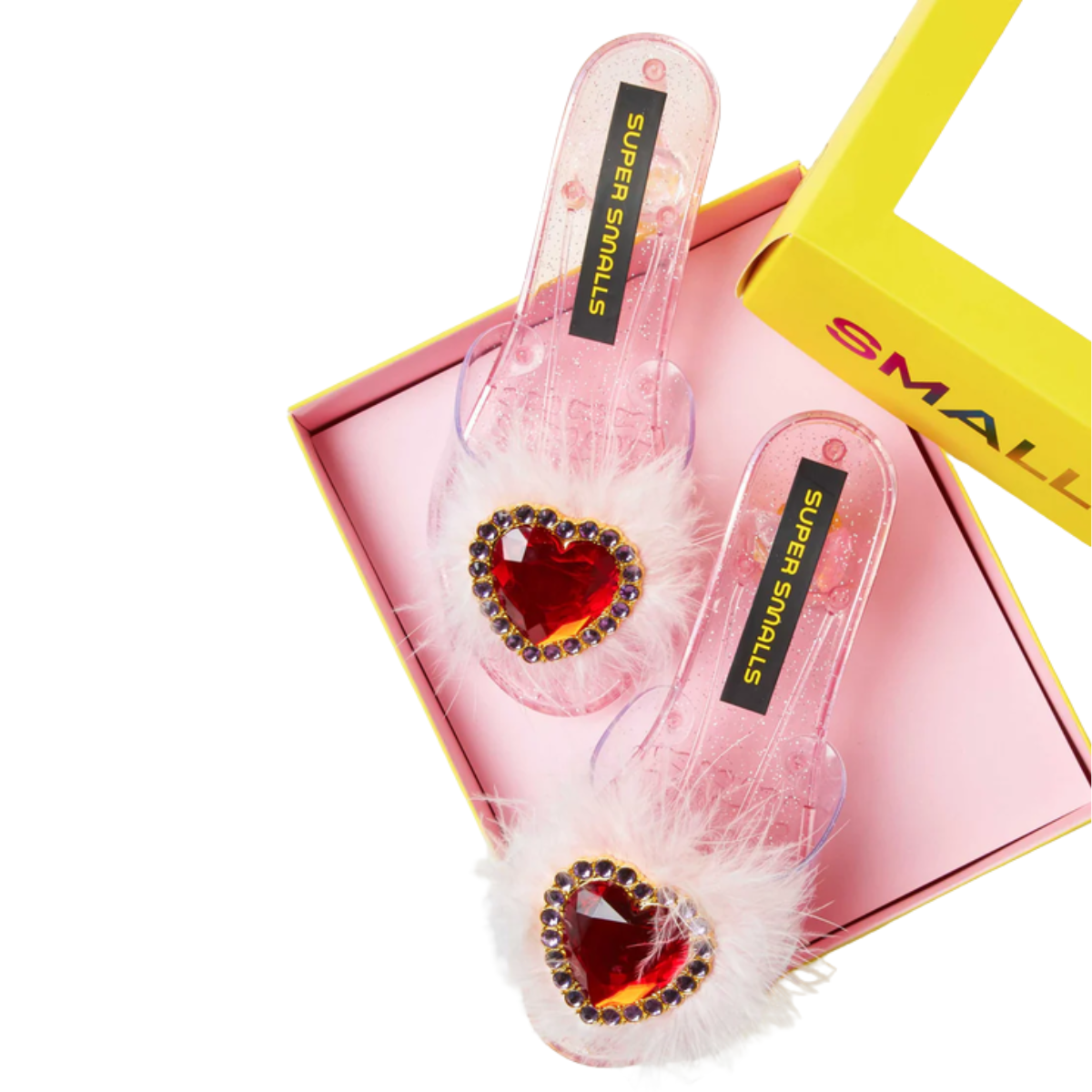 A pair of pink transparent slide sandals with white feather trim and a red heart-shaped rhinestone on the toe, displayed in a pink box with a yellow lid. The insoles read 'SUPER SMALLS'.