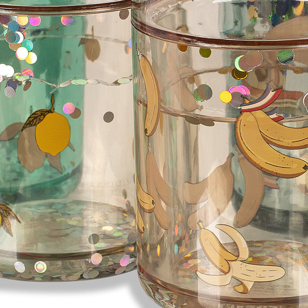 A close-up of several clear glass tumblers decorated with fruit illustrations and iridescent confetti. The front tumbler features bananas, peaches, and colorful glitter accents, with other tumblers in the background showing different decorative designs.
