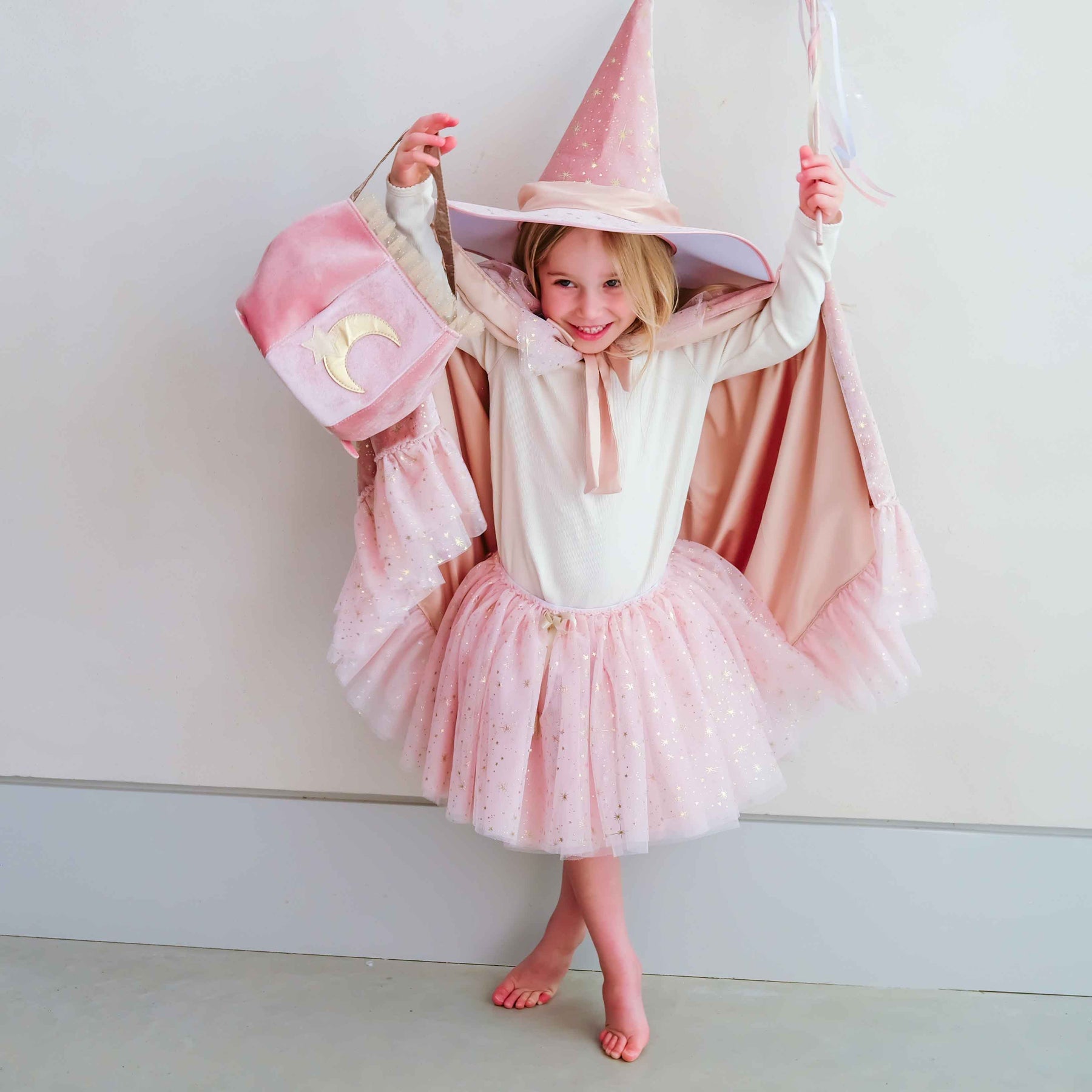A young girl posing in a pink and white wizard costume, featuring a tall pointed hat, a tutu skirt, and a cape with a moon emblem, set against a plain light gray wall.