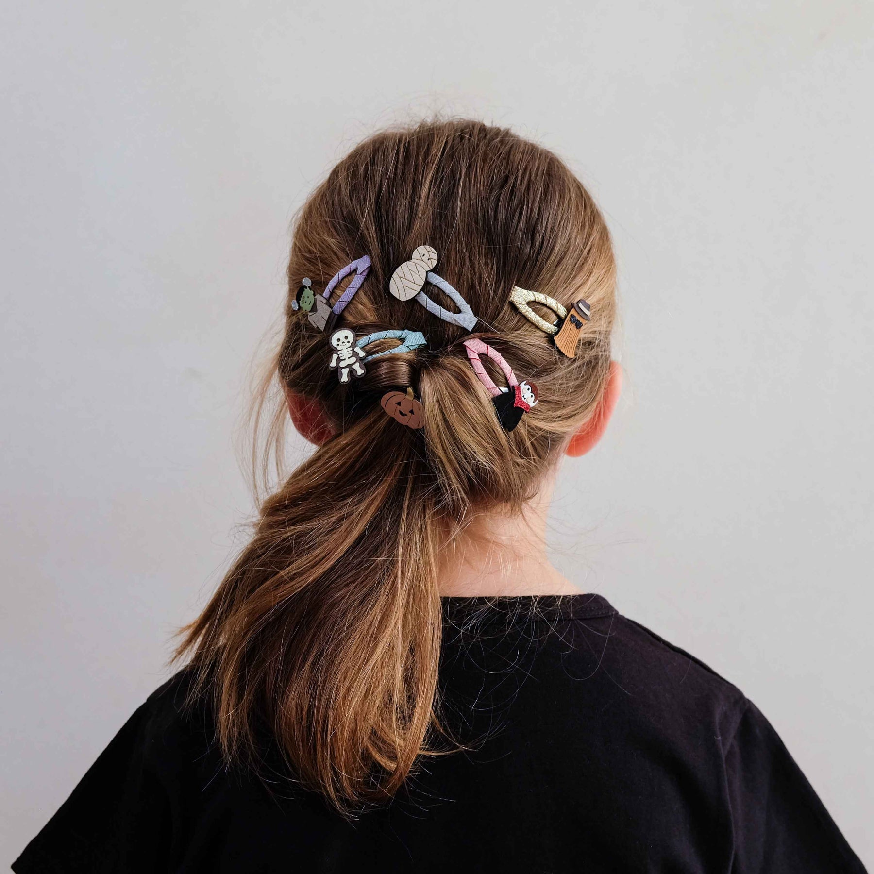 A person shown from the back wearing a black top, with light brown hair tied in a low ponytail and secured with multiple pastel-colored hair clips against a plain light gray wall.