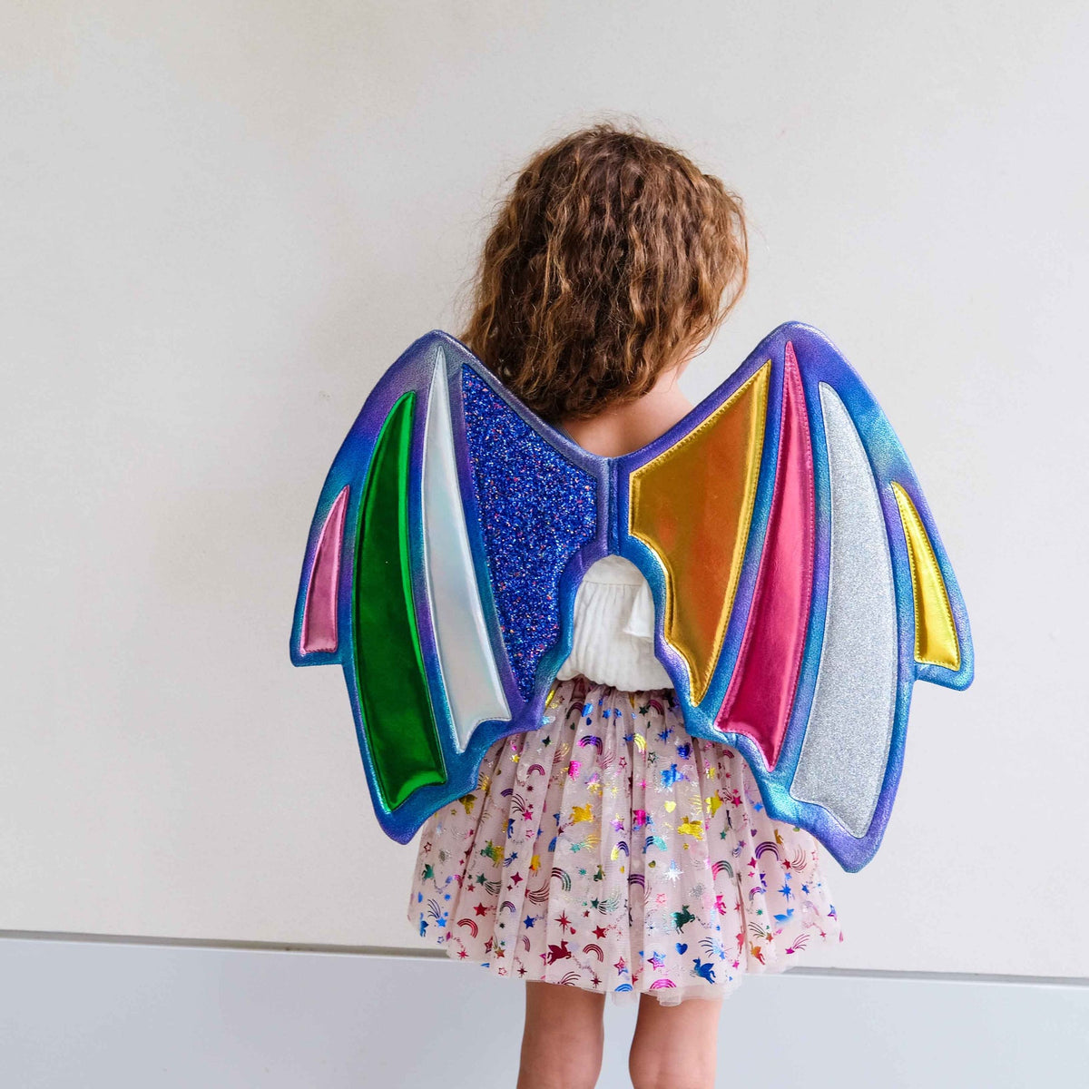 A child viewed from behind wearing a colorful fairy wing costume accessory with glittery blue, pink, green, yellow, orange, and silver panels, paired with a light pink floral skirt, against a plain white wall.