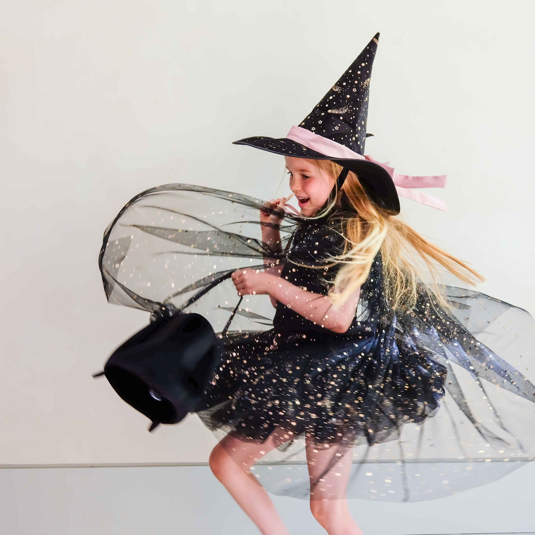 A young girl wearing a black witch costume with a glittery dress, pointed hat, sheer veil, and a black bucket bag, captured in motion against a plain light background.