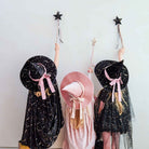 Three children wearing witch costumes stand against a plain light wall, each holding a black star wand. The child on the left wears a black dress with gold speckles and a large black hat with a pink ribbon.
