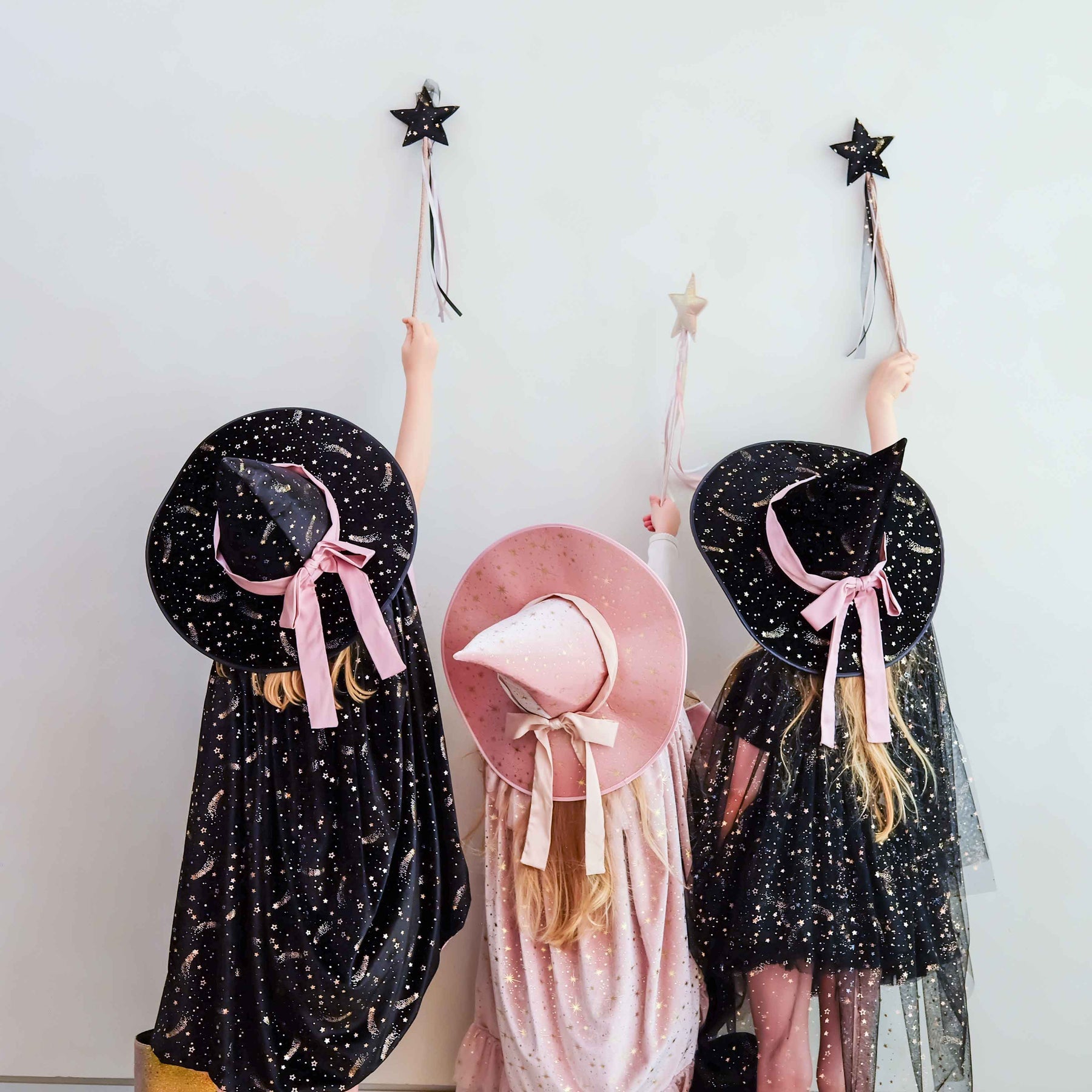 Three children wearing witch costumes stand against a plain light wall, each holding a black star wand. The child on the left wears a black dress with gold speckles and a large black hat with a pink ribbon.