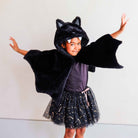 A young child wearing a black plush bat costume hood with attached wings, a black tulle skirt with silver star print, and a dark purple top, posing with arms outstretched against a plain light gray wall.