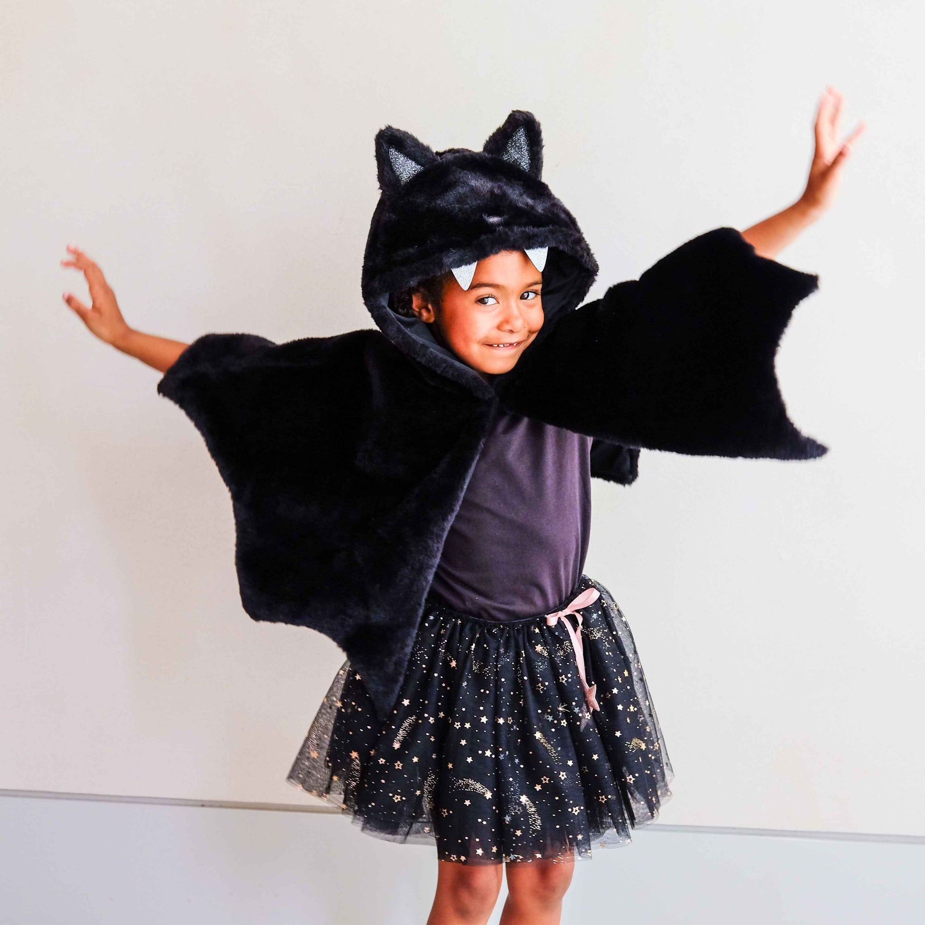 A young child wearing a black plush bat costume hood with attached wings, a black tulle skirt with silver star print, and a dark purple top, posing with arms outstretched against a plain light gray wall.