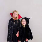 Two young girls wearing costume capes, standing against a plain light gray wall. The girl on the left wears a black cape with silver star patterns and a red inner collar, while the girl on the right wears a black plush cat-ear cape.