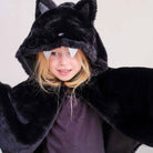 A child wearing a plush black bat costume hood with pointed ears, silver glitter bat wings attached to the hood, and a dark purple shirt underneath, photographed against a plain light background.