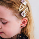 A close-up of a young girl's head and ear, showcasing two gold hair claw clips with colorful pastel house charms and matching small stud earrings.