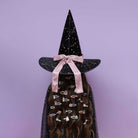 A person wearing a tall black witch hat with gold speckles, a pink satin bow, and a matching black cape covered in colorful hair clips, set against a solid lavender background.