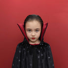 A young girl modeling a vampire costume cape with a black starry exterior and a deep red inner lining, set against a solid red background.
