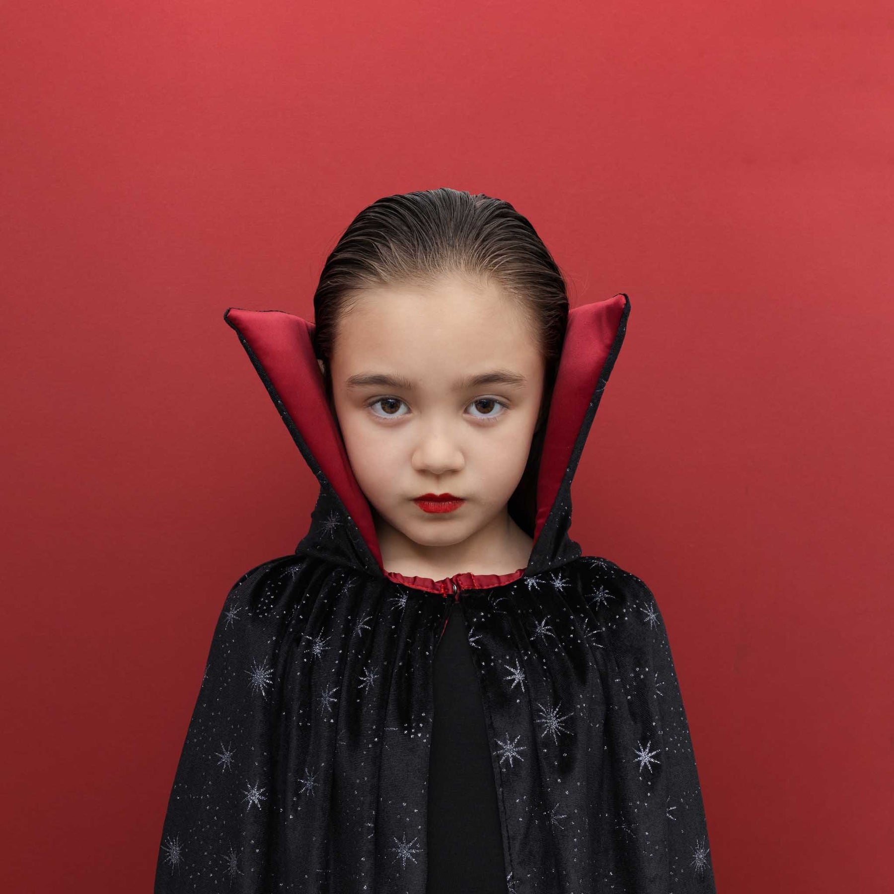 A young girl modeling a vampire costume cape with a black starry exterior and a deep red inner lining, set against a solid red background.