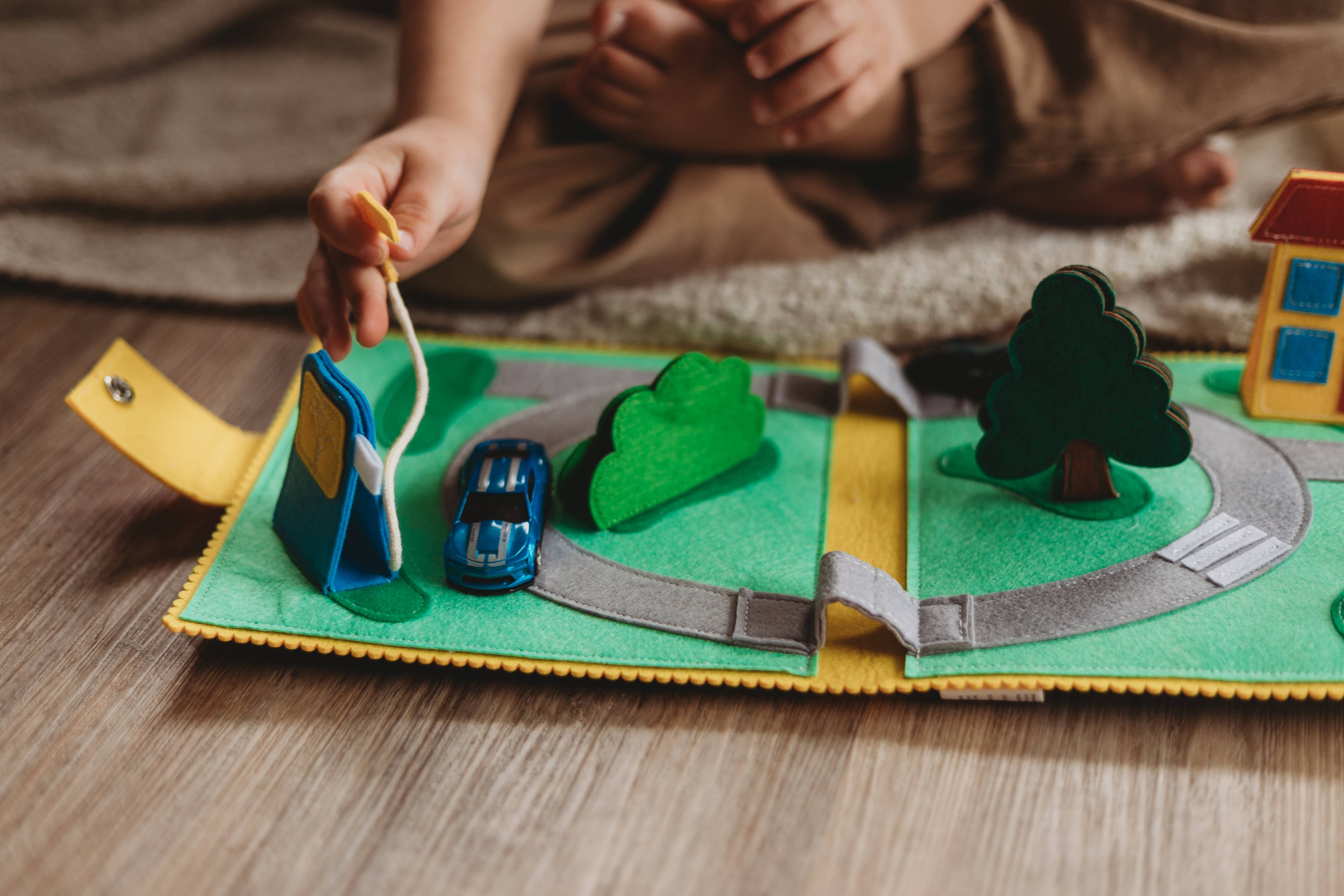 A close-up of a child's hands playing with a felt road play mat on a wooden floor. The green felt mat features a winding gray road, a blue toy car, a green tree, a yellow sign, and a small house.