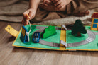 A close-up of a child's hands playing with a felt road play mat on a wooden floor. The green felt mat features a winding gray road, a blue toy car, a green tree, a yellow sign, and a small house.