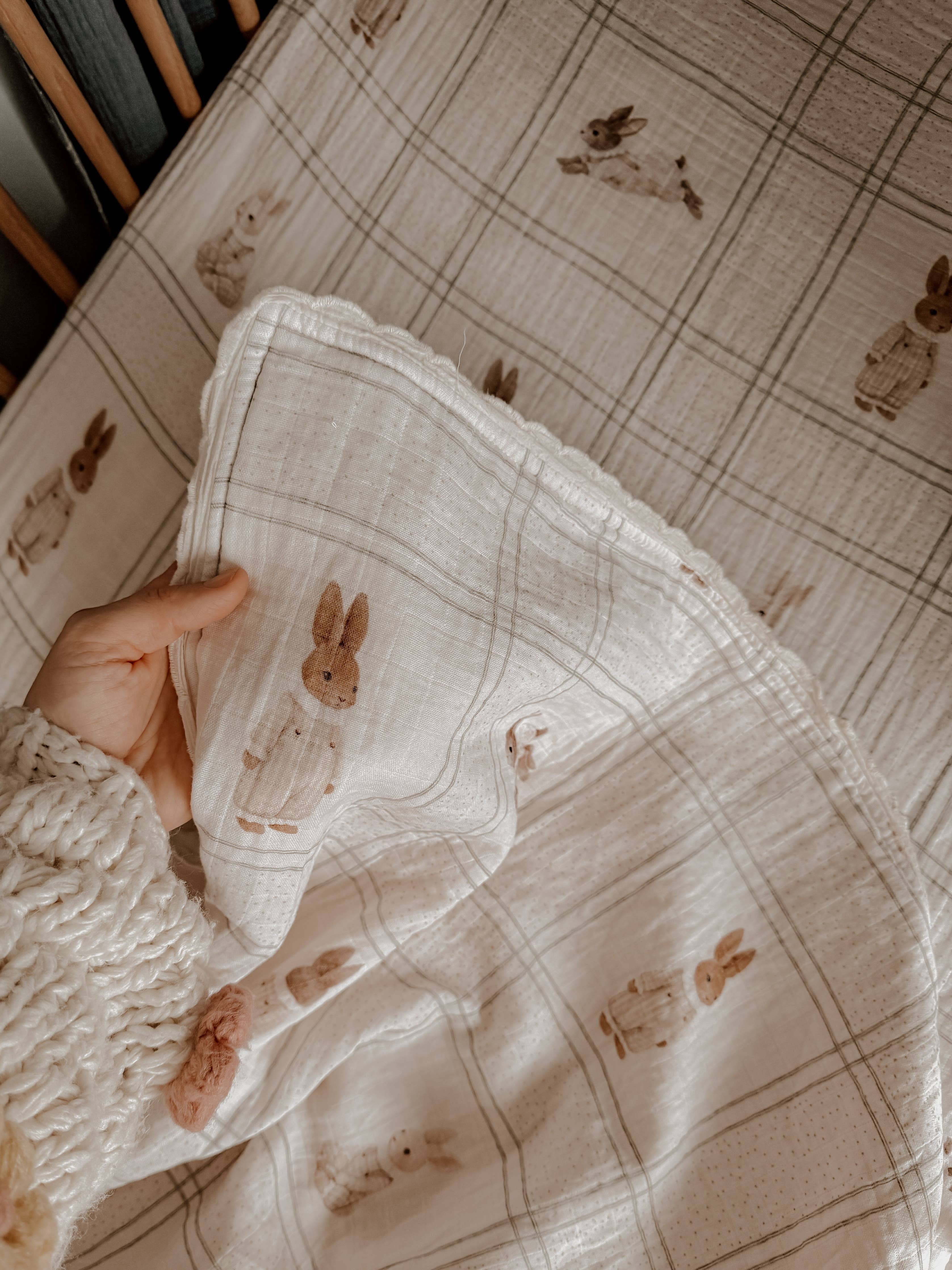 A close-up of a hand holding a cream-colored muslin blanket with a brown bunny print, set against a matching plaid sheet.