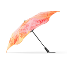 A compact folding umbrella shown open on a plain white background. The canopy has a warm gradient pattern shifting from orange to pink, with a subtle floral or abstract print, while the handle and shaft are black.