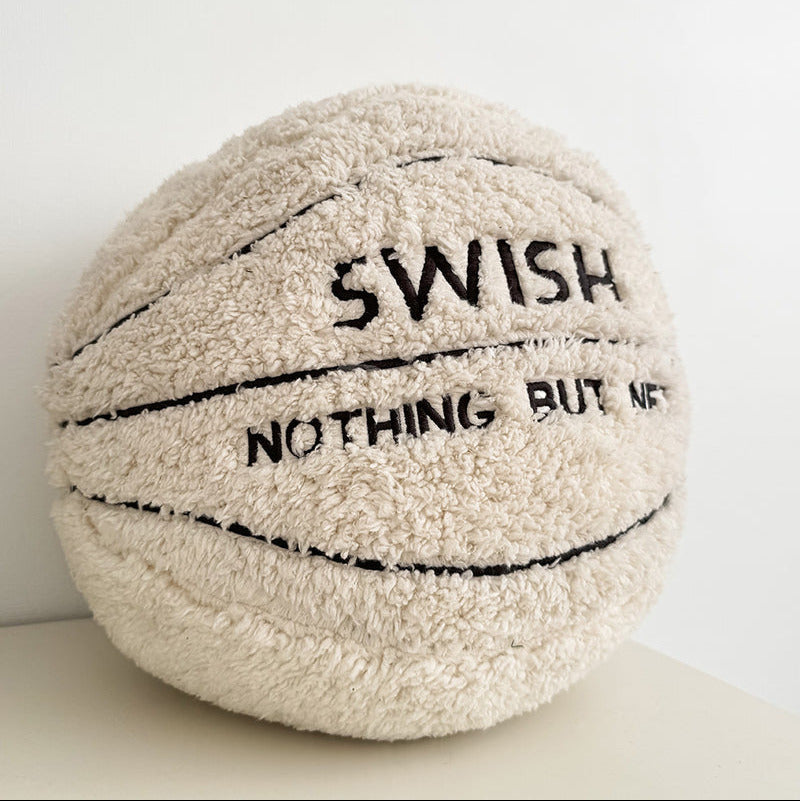 A plush, basketball-shaped throw pillow in cream-colored sherpa fabric with black embroidered lines and text reading 'SWISH NOTHING BUT NAF'.