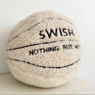 A plush, basketball-shaped throw pillow in cream-colored sherpa fabric with black embroidered lines and text reading 'SWISH NOTHING BUT NAF'.