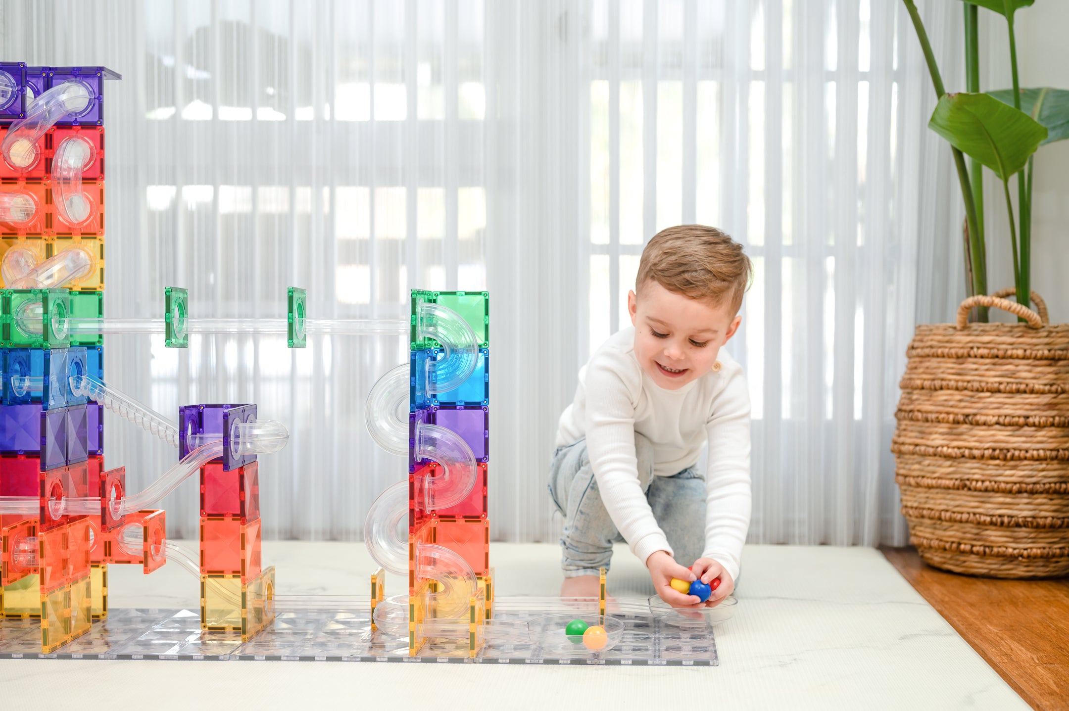 A young child in a white long-sleeve shirt and light blue jeans is kneeling on a white floor, playing with a colorful translucent magnetic marble run set. The scene is set in a bright, modern room with sheer white curtains, a green plant, and a woven basket.