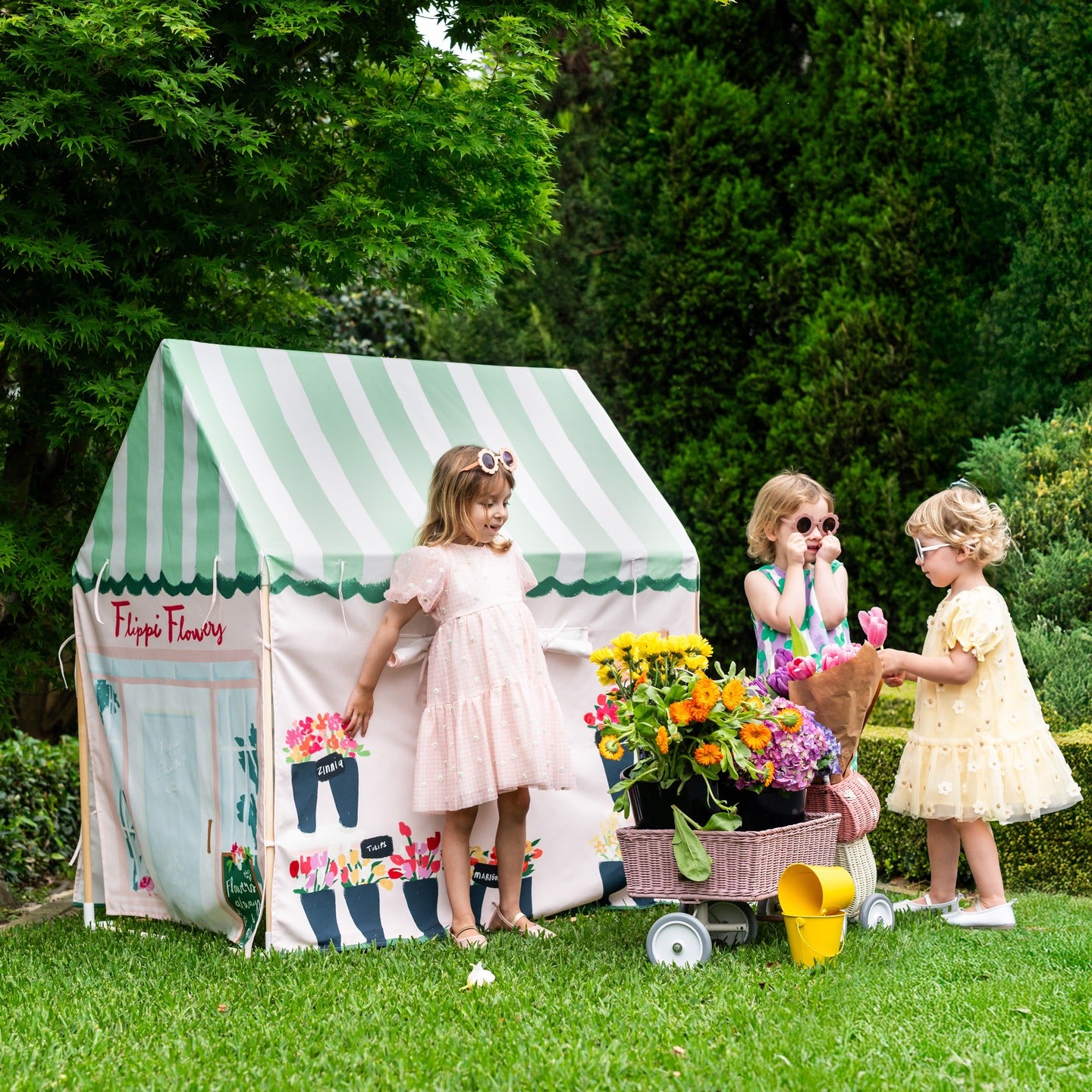 Three young girls play outdoors in a grassy garden with a small Flippi striped play tent, a flower cart, and a yellow watering can. The play tent is printed with floral designs and the text 'Flippi Flower'.