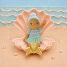 A plush mermaid doll with brown skin, blue hair, and a light blue and green dress sits inside a large pink seashell on a sandy beach set with blue ocean waves and pearl-like beads.