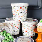 Four white Halloween-themed ice cream tubs with lids, decorated with colorful spooky illustrations like bats, ghosts, pumpkins, and candy corn, arranged on a black surface with decorative props including green-striped lollipops and a cupcake.