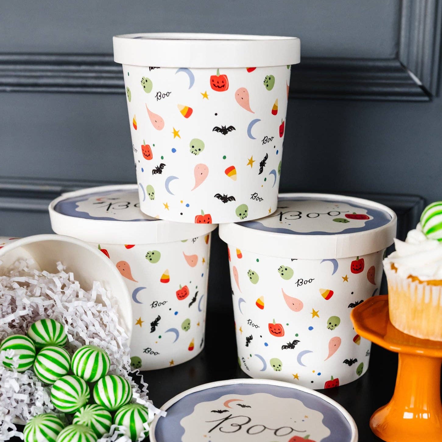 Four white Halloween-themed ice cream tubs with lids, decorated with colorful spooky illustrations like bats, ghosts, pumpkins, and candy corn, arranged on a black surface with decorative props including green-striped lollipops and a cupcake.