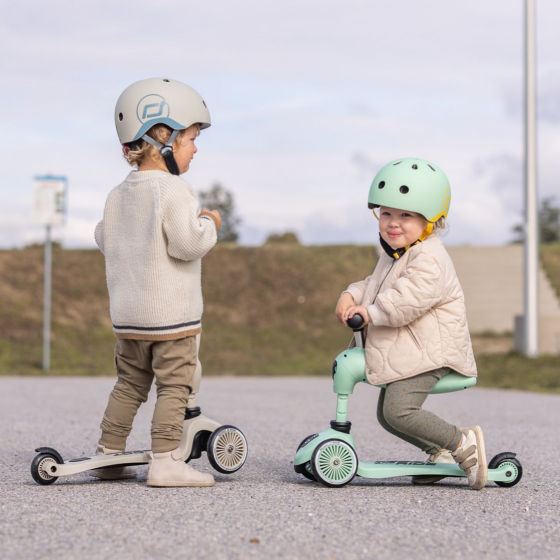Two children on scoot n ride scooters wearing helmets in an outdoor setting