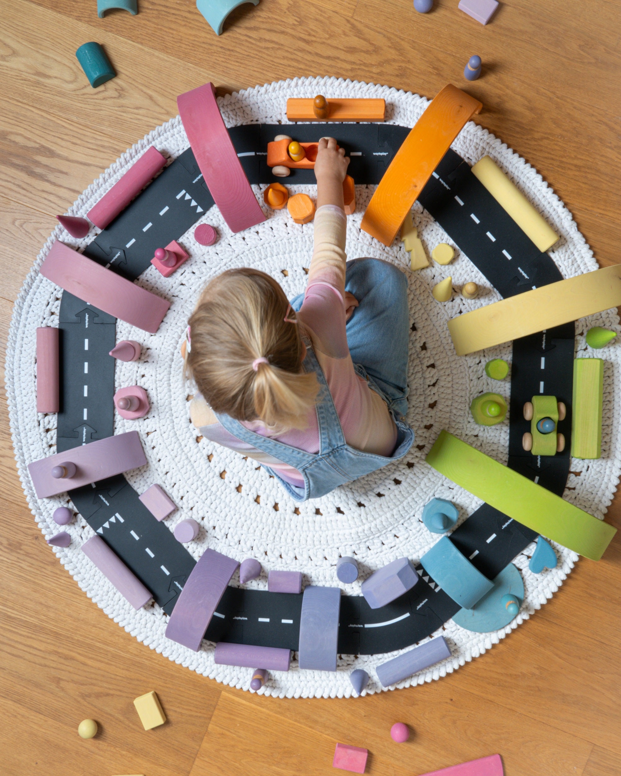 A child sits on a round white crocheted rug surrounded by a colorful wooden toy road set with black roads, pink and orange bridges, yellow and green sections, and small toy cars, on a wooden floor.