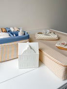 A styled baby changing table setup featuring a striped changing pad, a blue fabric diaper basket filled with baby items, a white ceramic house-shaped container, and a patterned tissue pouch.