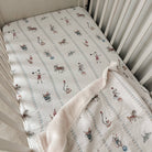 A baby crib mattress fitted sheet with a white background and a playful print of vintage carousel horses and riders in muted red, blue, and brown, with a matching plush cream blanket draped over the corner.