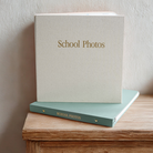 A hardcover photo album titled 'School Photos' in gold lettering, standing upright on top of another matching album on a wooden table against a textured beige wall.