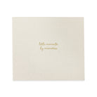 A square cream-colored fabric keepsake album or photo book with gold foil text reading 'little moments by memories' in the center, shown on a plain white background.