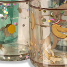 A close-up of several clear glass tumblers decorated with fruit illustrations and iridescent confetti. The front tumbler features bananas, peaches, and colorful glitter accents, with other tumblers in the background showing different decorative designs.